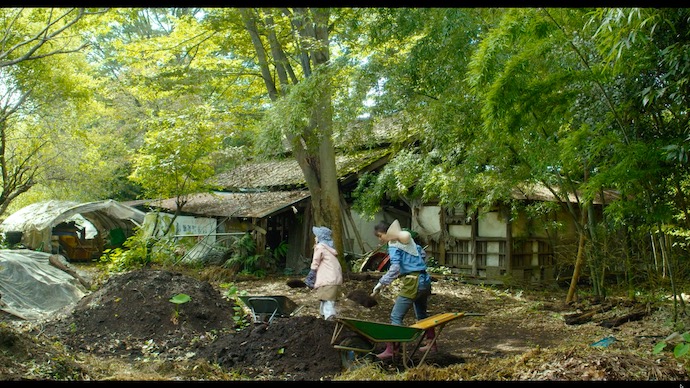 生い茂る木々に囲まれた古い小屋の前で、大きなスコップを手に持ち、作業をする女性2人